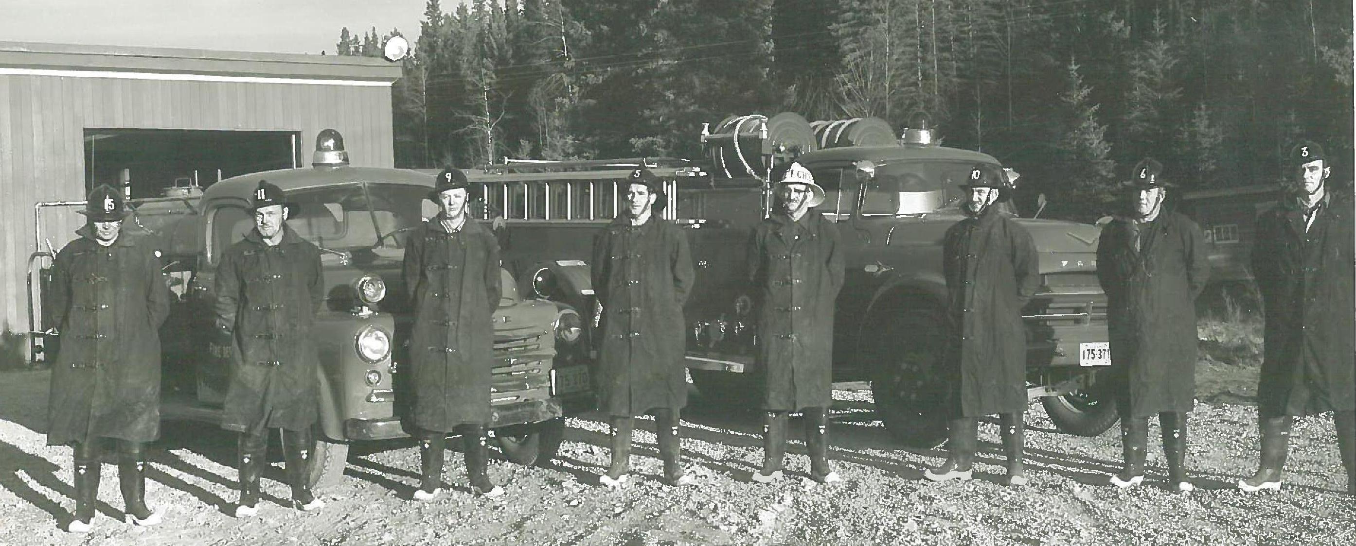 Old historical photo of 8 firefighters in front of some old fire trucks.